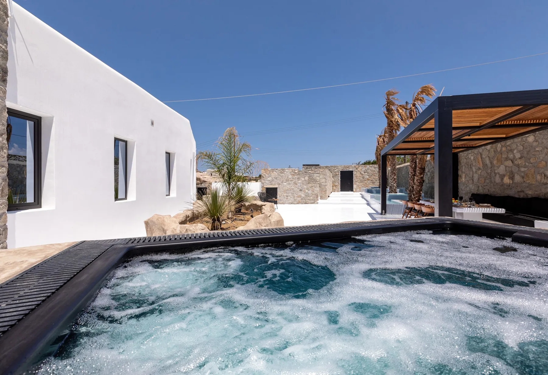 Una piscina exterior con burbujas está junto a un edificio blanco y una zona de estar sombreada.