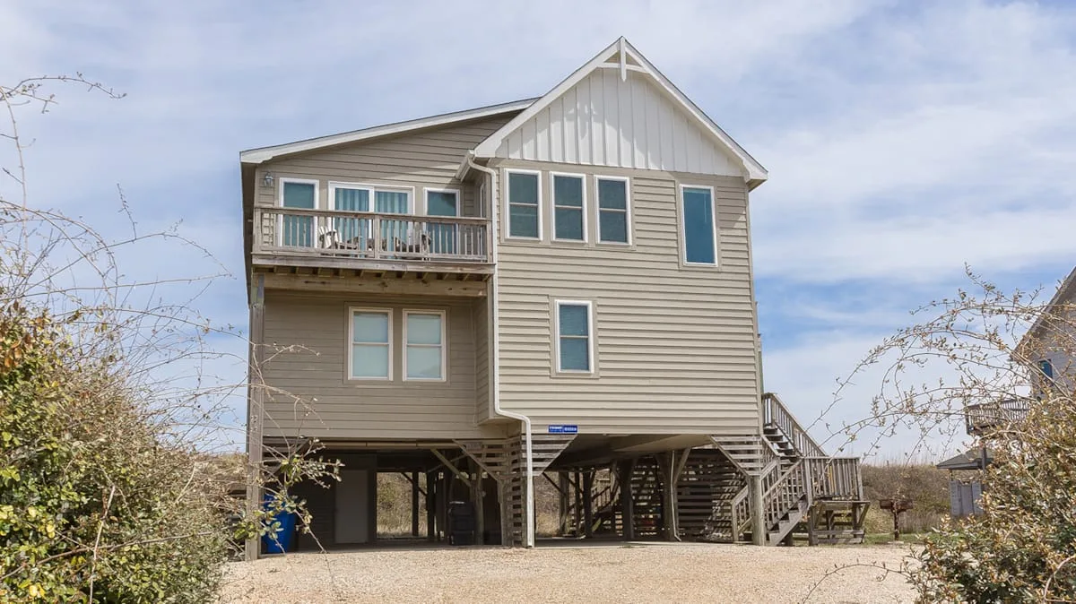 A raised house with exterior stairs stands among sparse trees beneath a cloudy sky.
