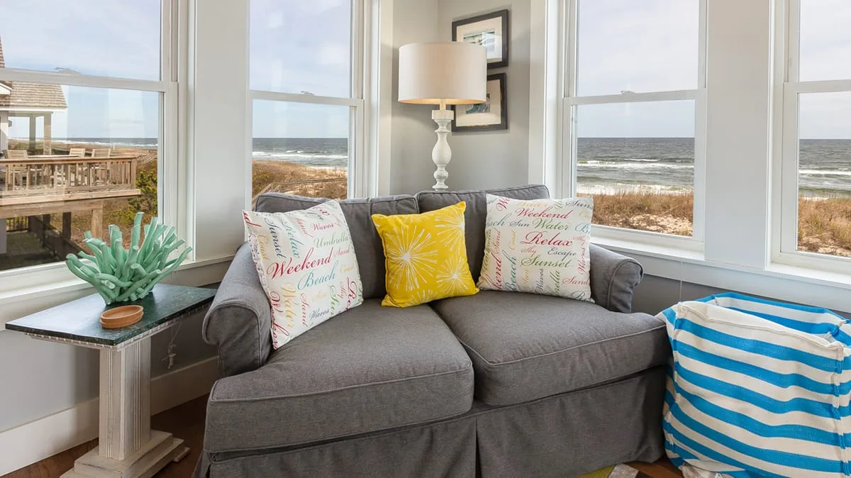 A living room with gray seating, colorful cushions, and large windows overlooking the ocean.