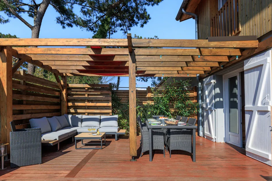 A wooden outdoor deck has seating beneath a pergola, with a dining table nearby.