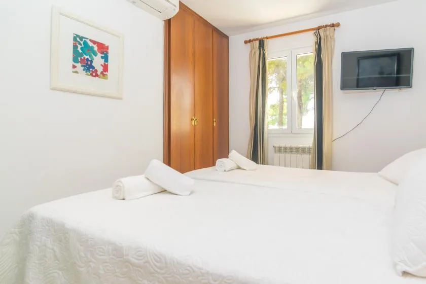 Un dormitorio luminoso con ropa de cama blanca, una ventana con cortinas y un televisor en la pared.