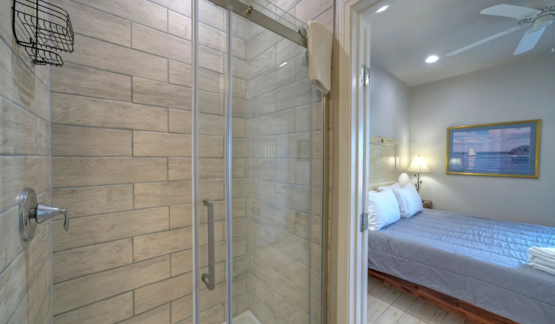 Una zona de ducha con azulejos se abre a un dormitorio con cama y arte enmarcado.