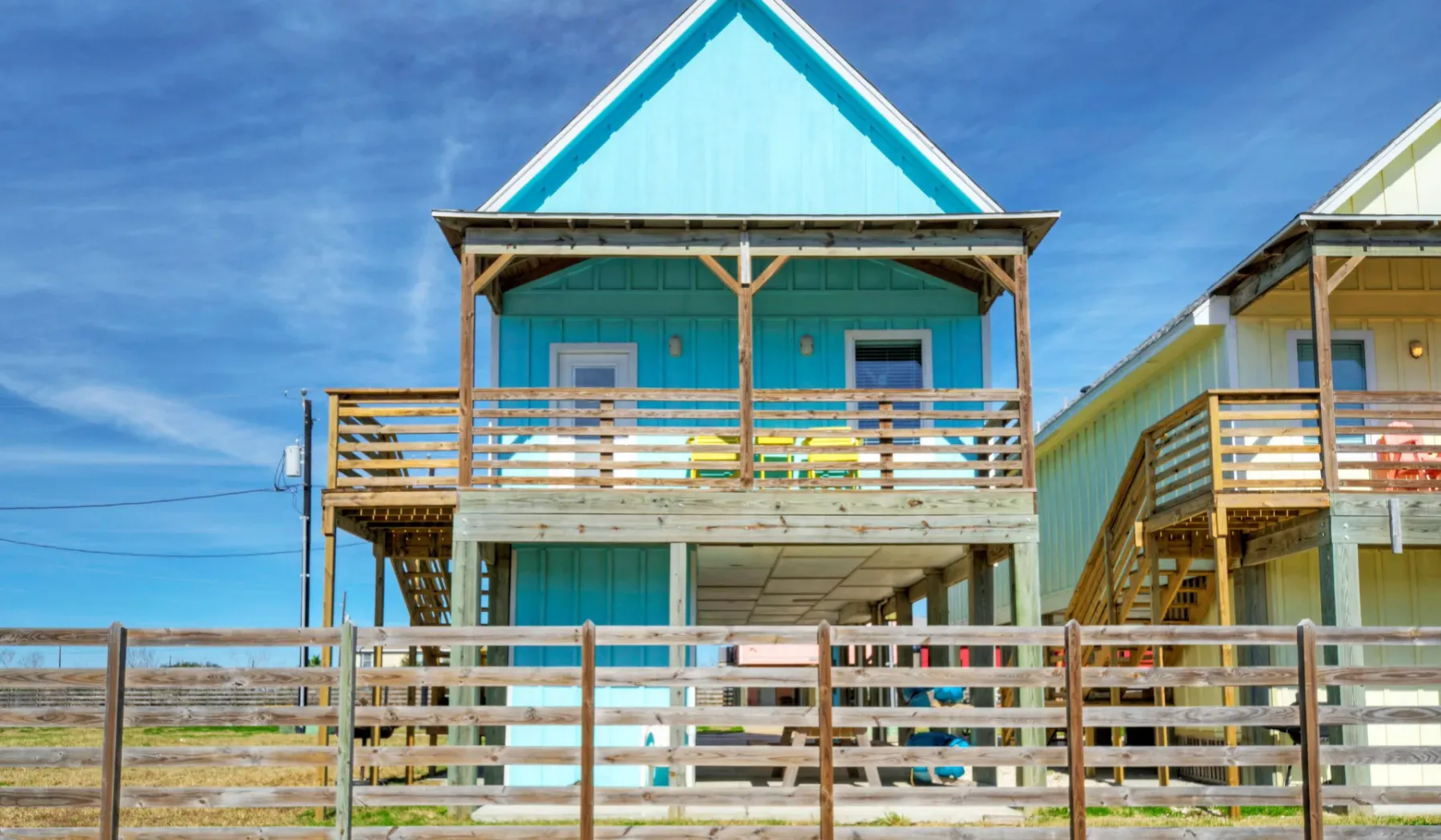 Una casa de playa turquesa se alza detrás de una cerca de madera bajo un cielo azul.