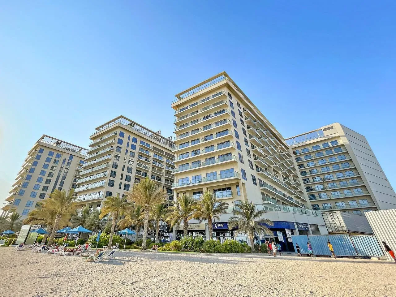 Un gran edificio de varios pisos se alza detrás de una playa de arena con palmeras.