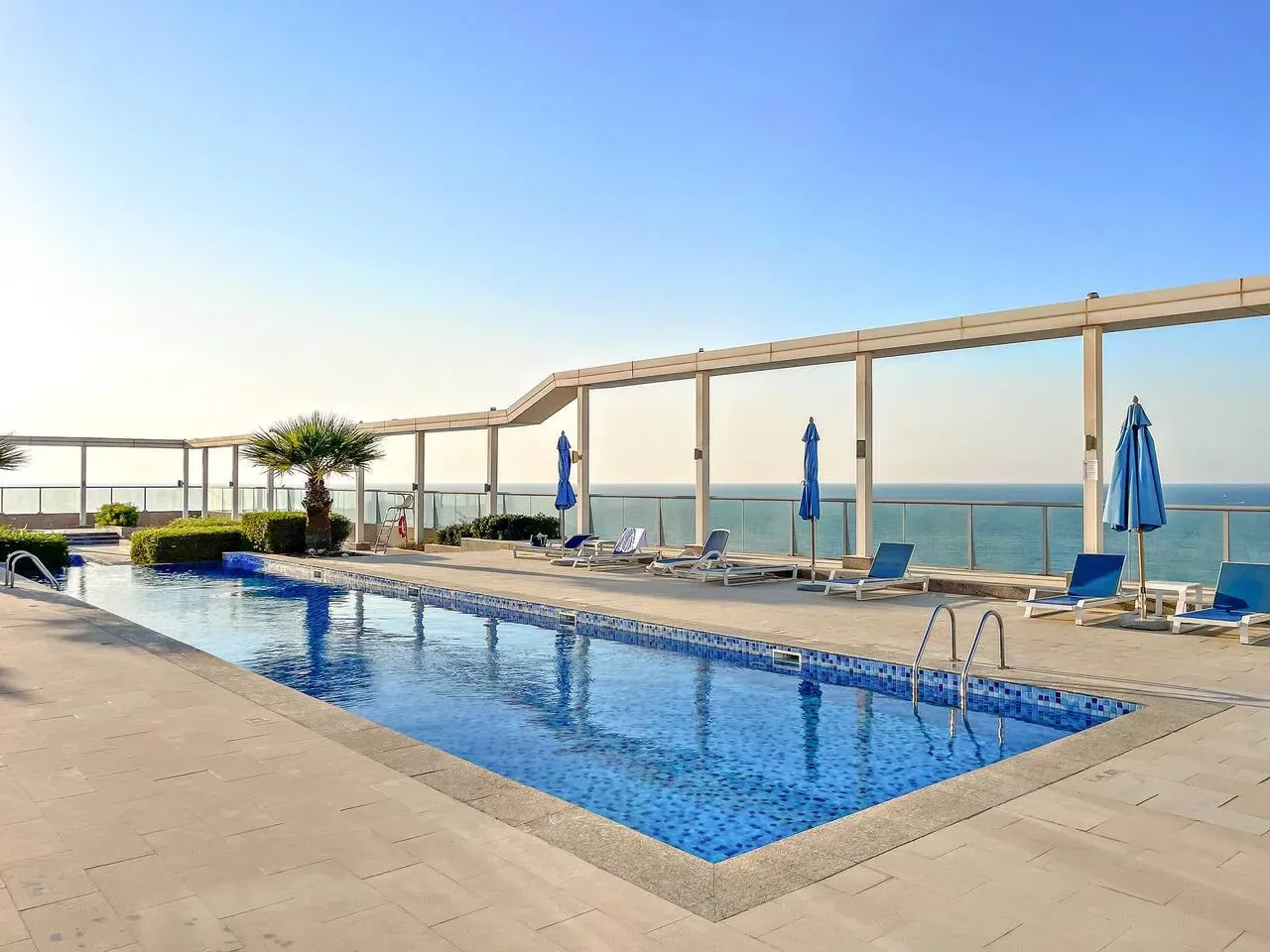 Una piscina al aire libre da al mar, con tumbonas y sombrillas azules cerca.
