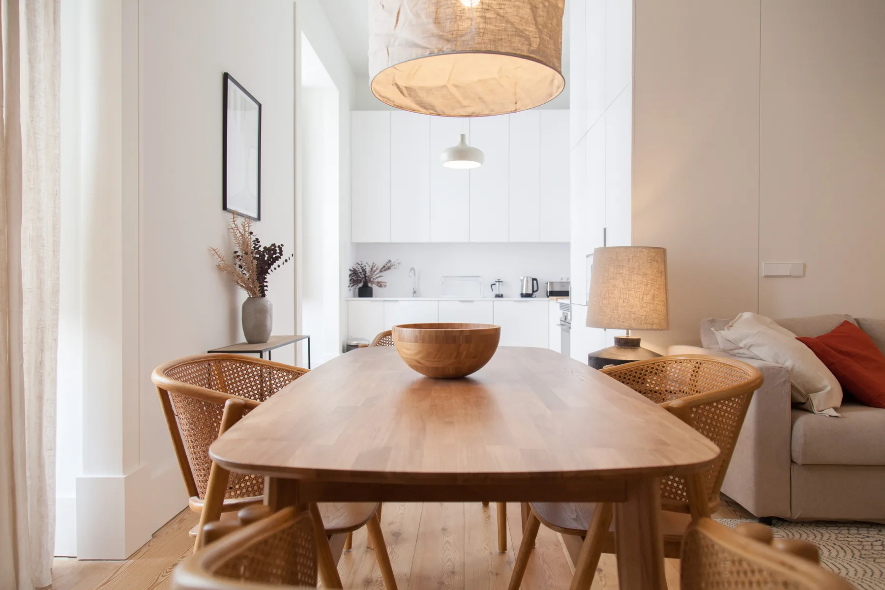 Un comedor luminoso da a una cocina blanca, con una mesa de madera y asientos tejidos.