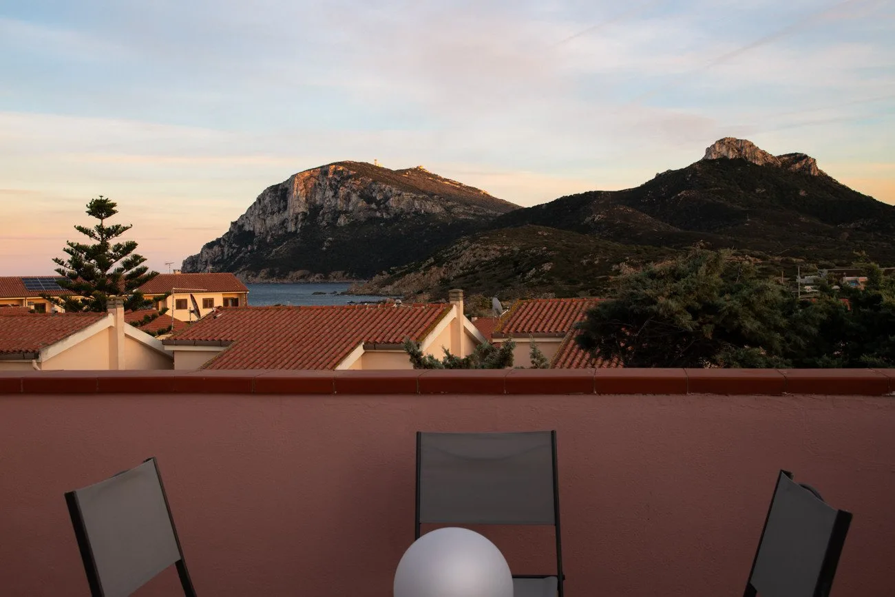 Una terraza en la azotea da a tejados de tejas rojas, el mar y montañas al atardecer.