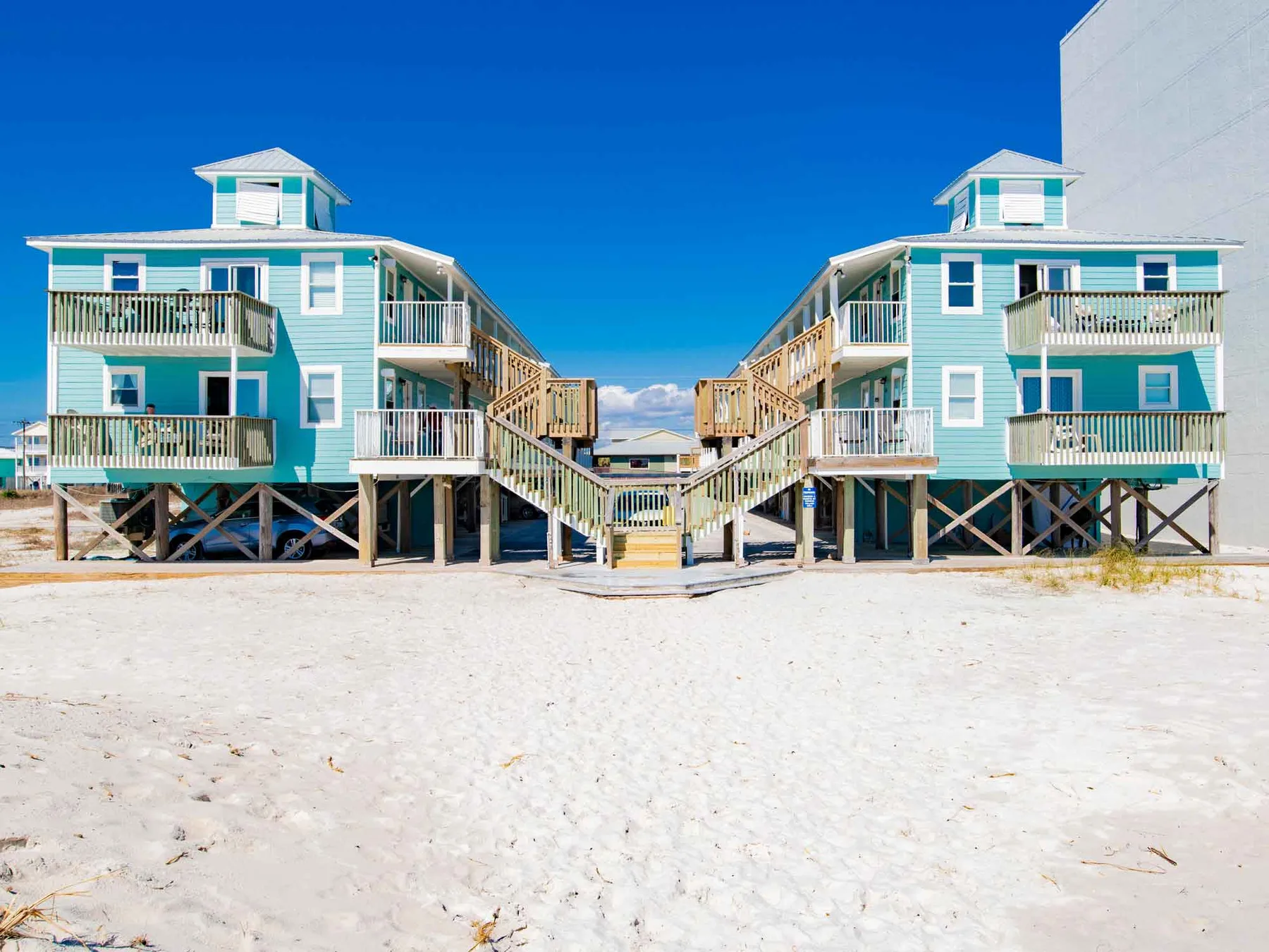 Dos casas de playa elevadas color turquesa se alzan sobre arena bajo un cielo azul despejado.