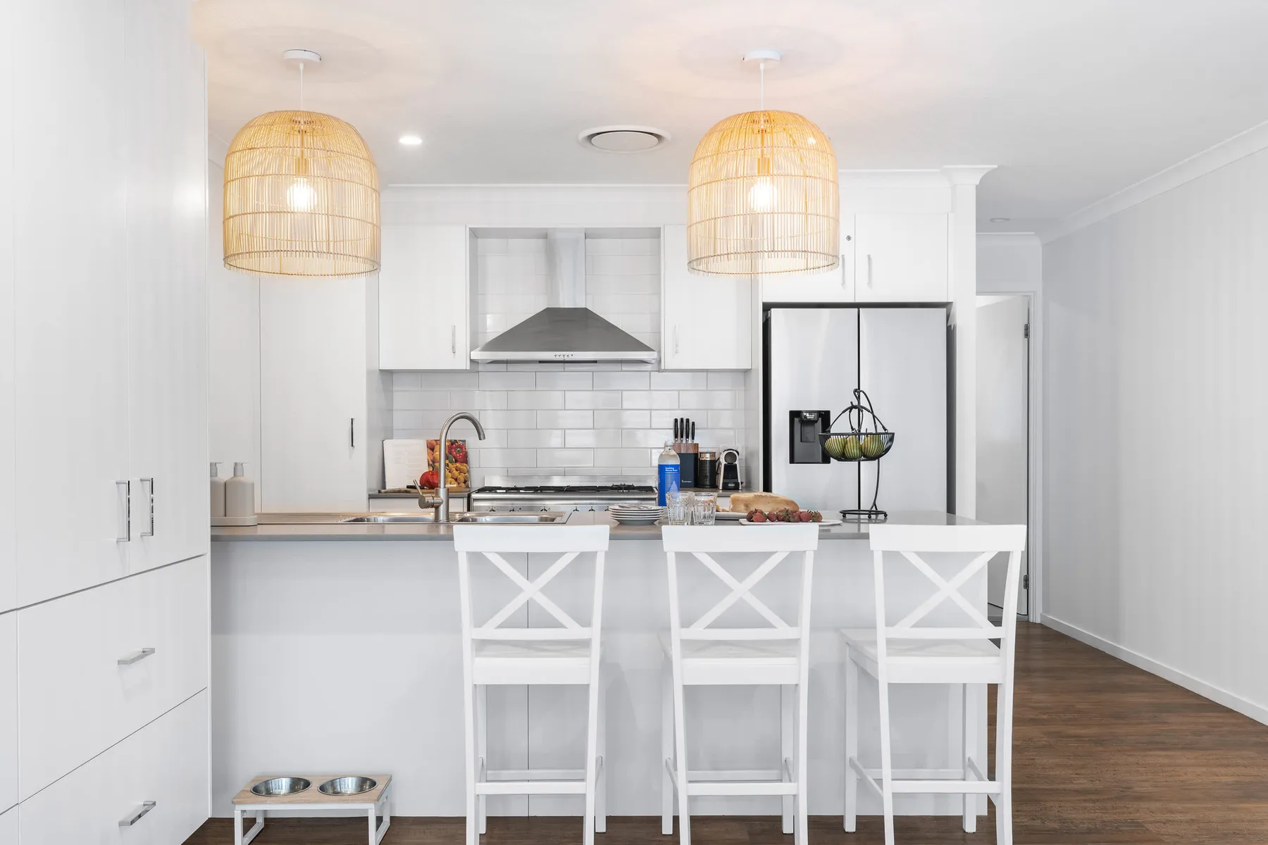 Una cocina blanca y luminosa con una isla y taburetes bajo lámparas colgantes tejidas.