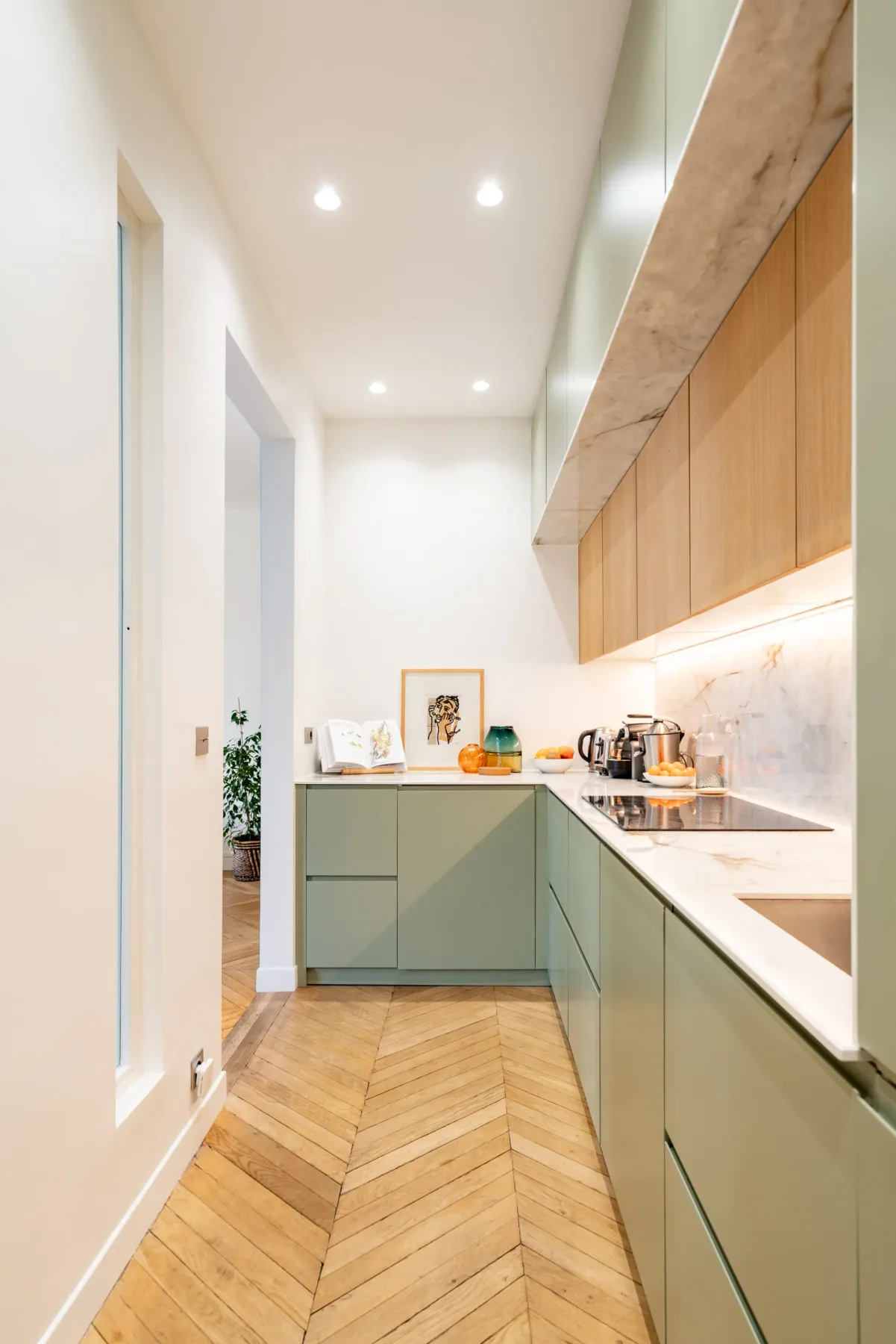 Una cocina alargada y luminosa con gabinetes verdes y piso con patrón de madera.