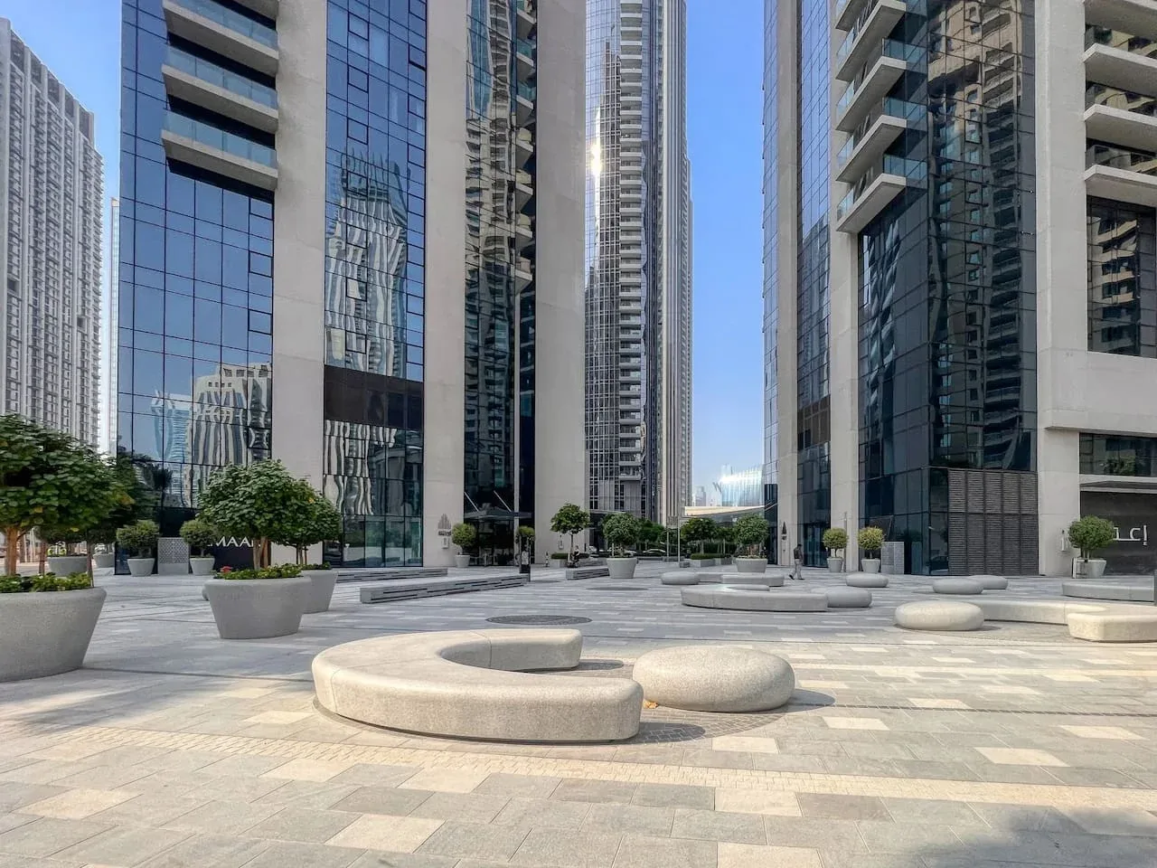 A paved plaza between tall glass buildings features circular stone seating and planters.