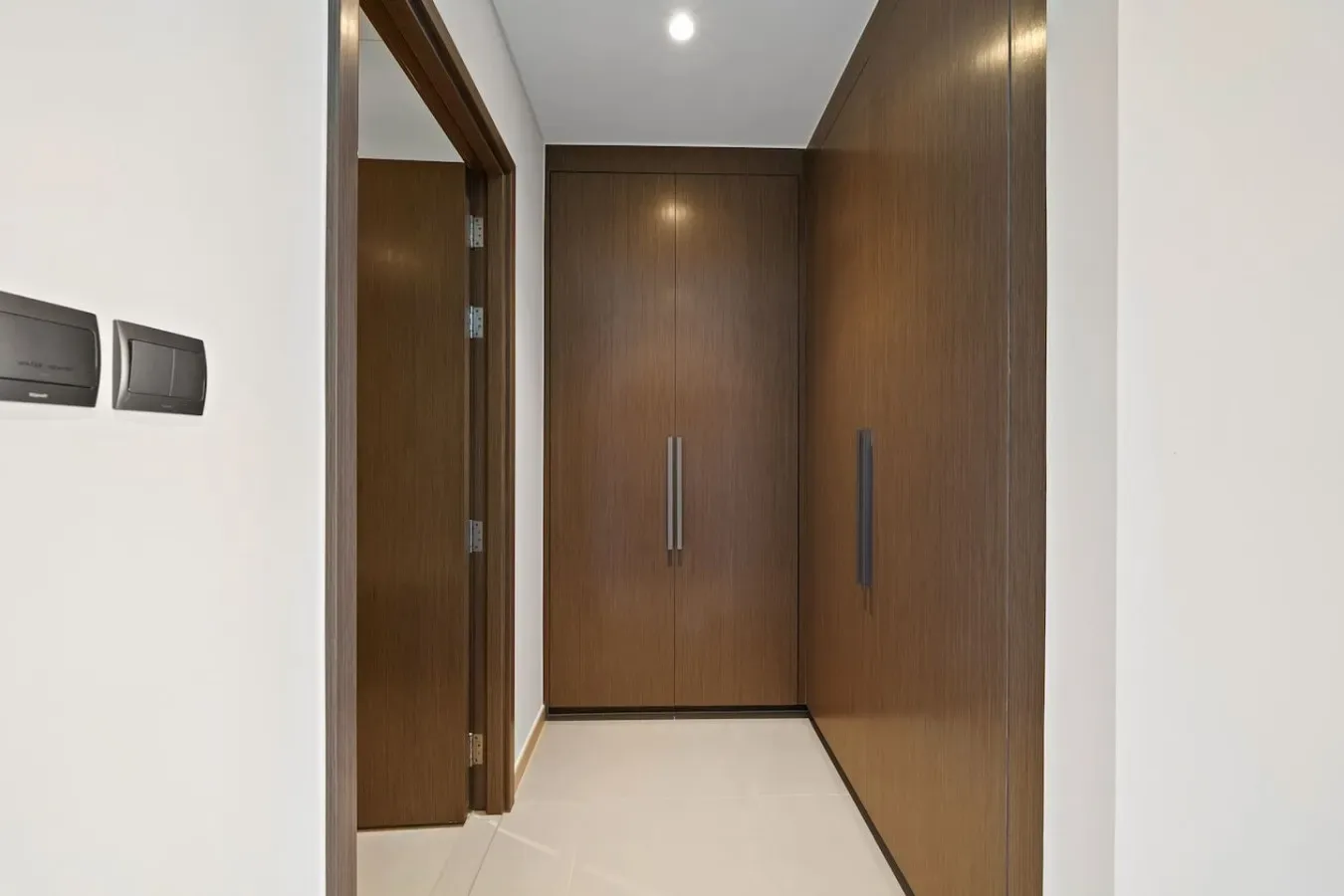 A narrow hallway with dark wooden doors and light tile flooring.