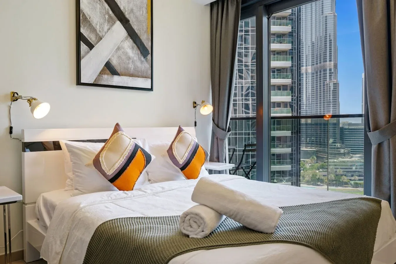 A bedroom with a neatly made bed and floor-to-ceiling windows overlooking nearby buildings.