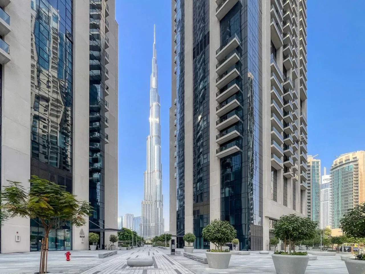 A paved plaza between tall buildings, with a slender tower visible in the distance.