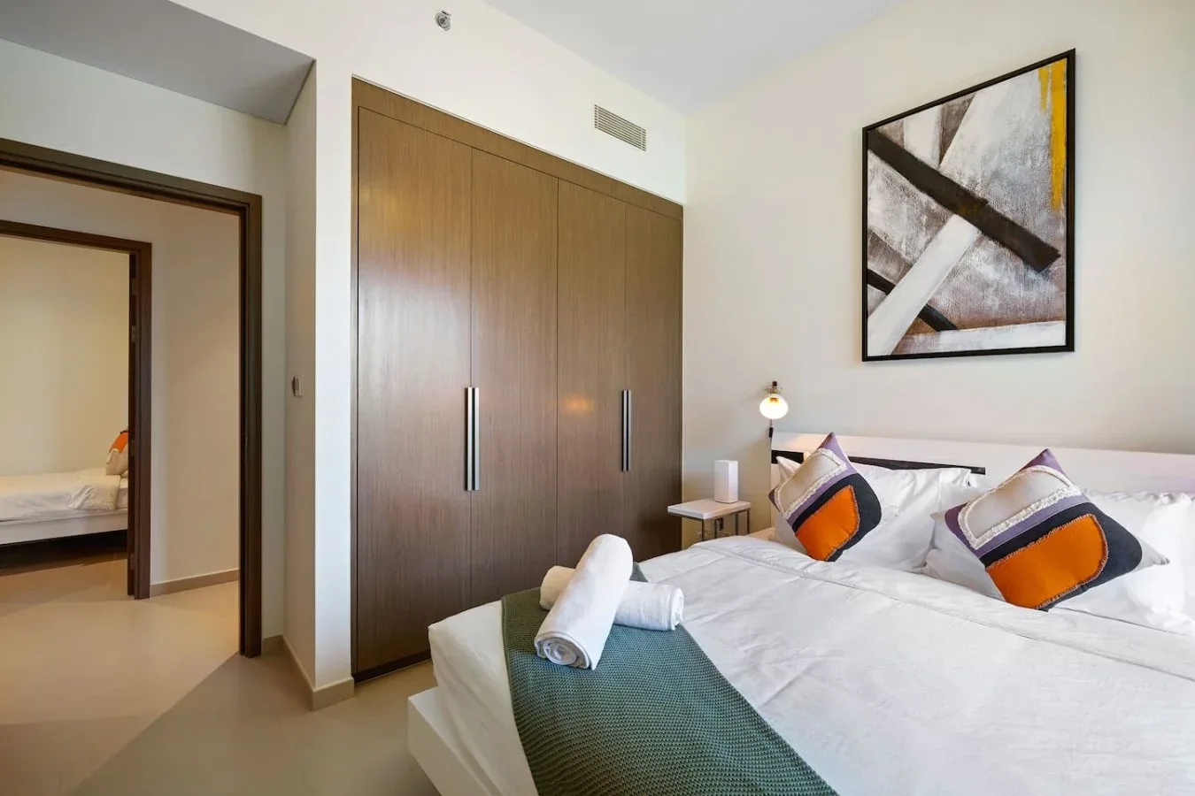 A bedroom with a neatly made bed, bedside lamp, and framed abstract artwork.