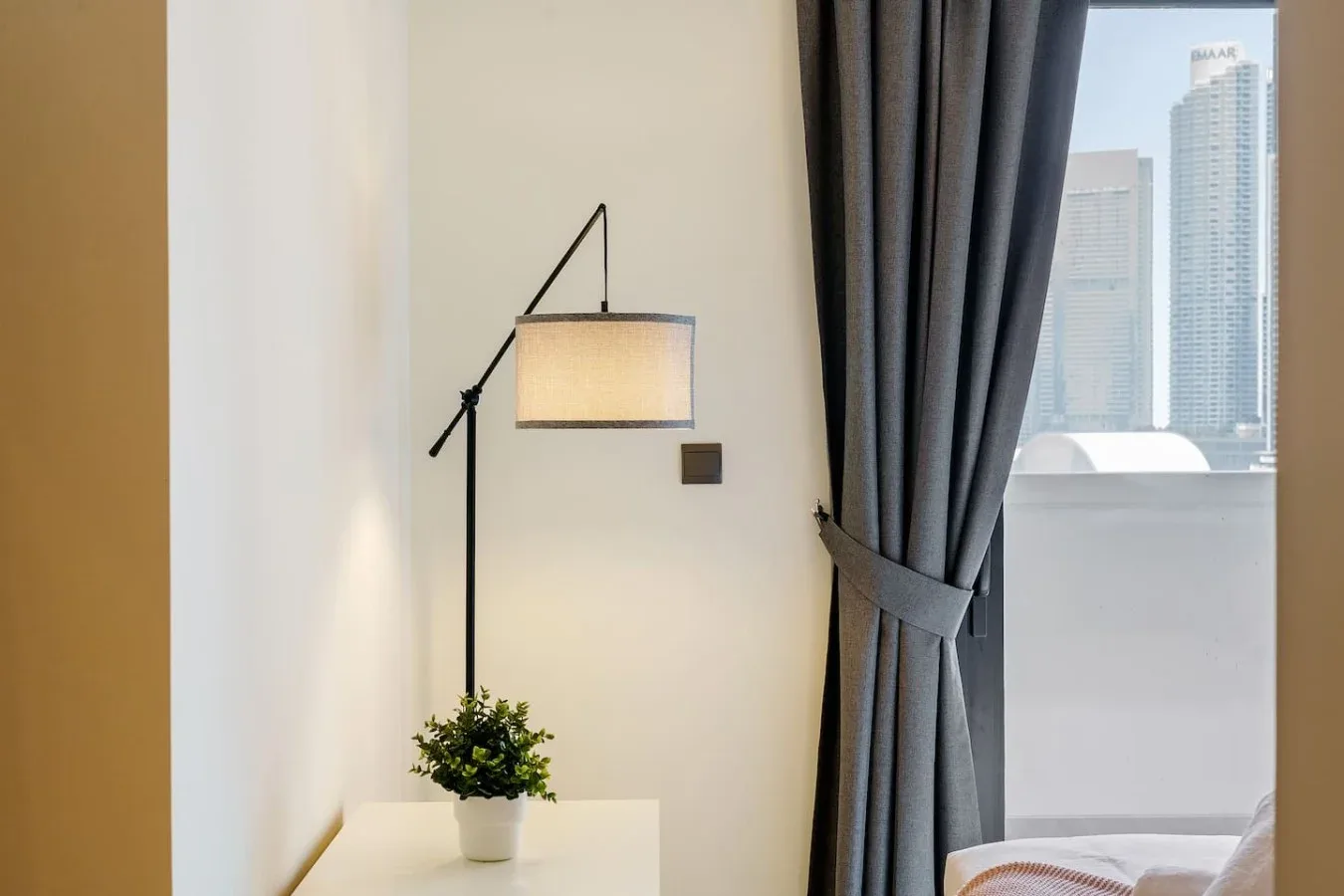 A bedroom corner with a floor lamp, a small plant, and a window overlooking city buildings.