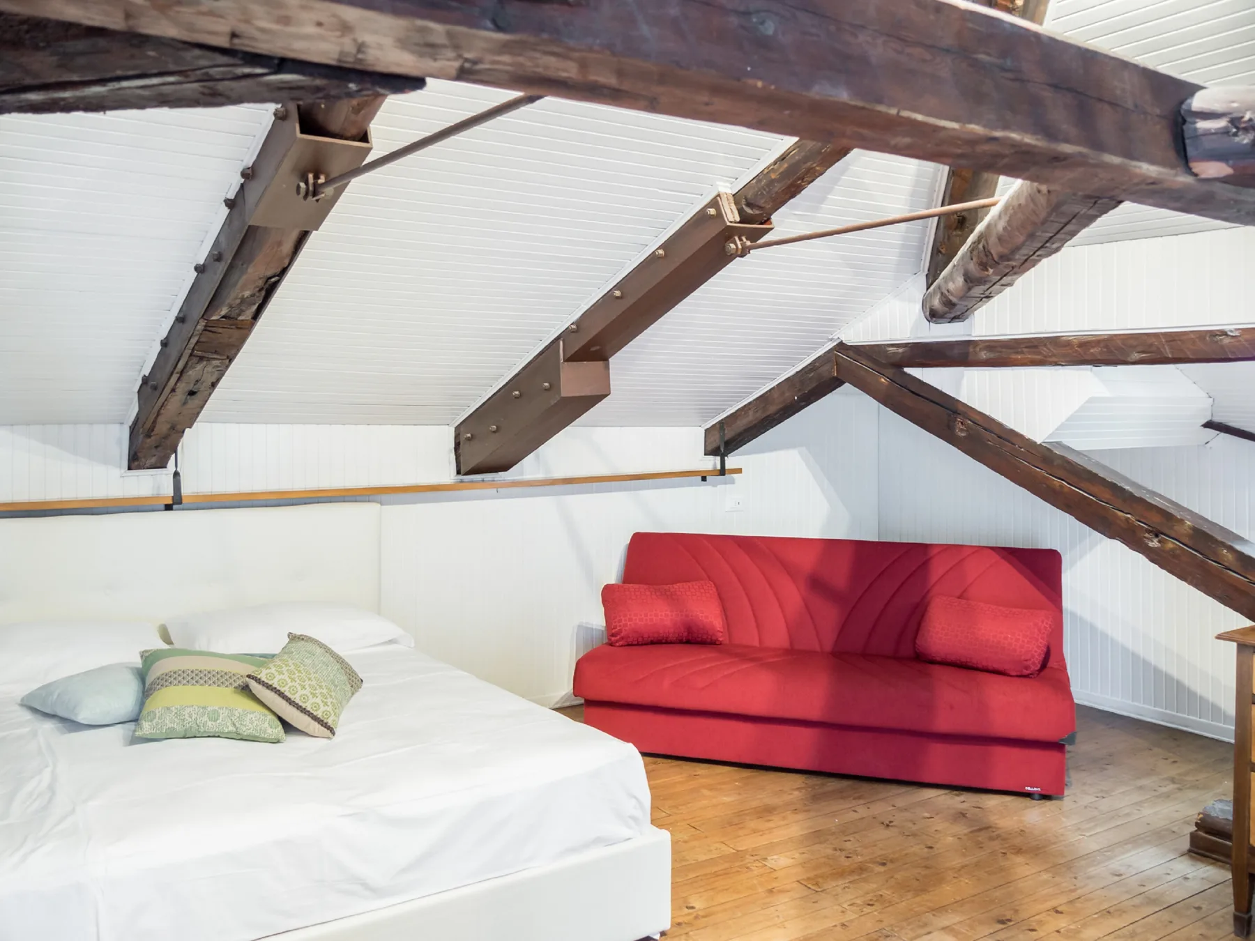 A bedroom with a red sofa beside a bed beneath exposed wooden beams.