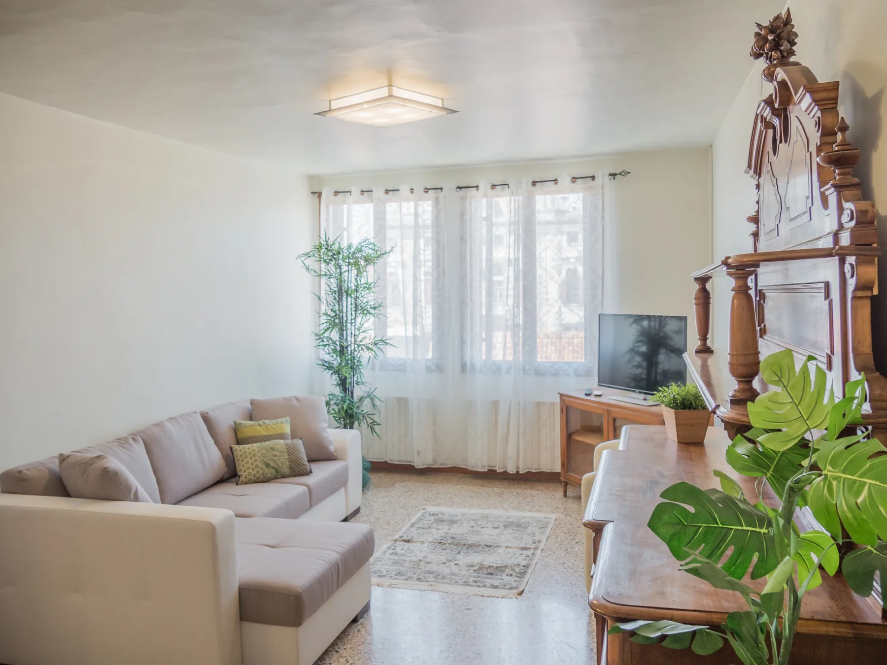 A bright living room with seating, a television, and sheer curtains over the windows.