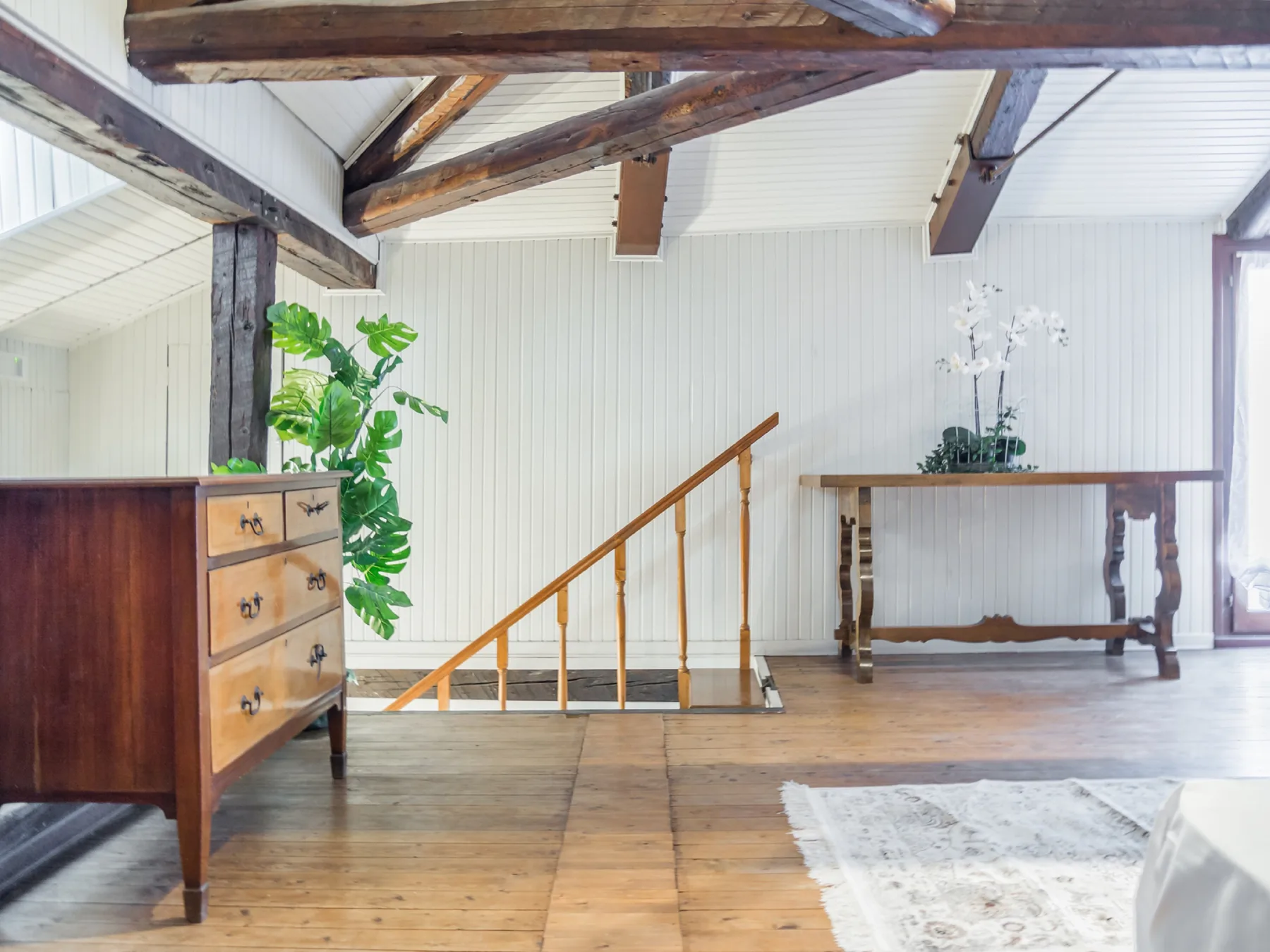 A bright room with exposed wooden beams, a staircase, and a table.