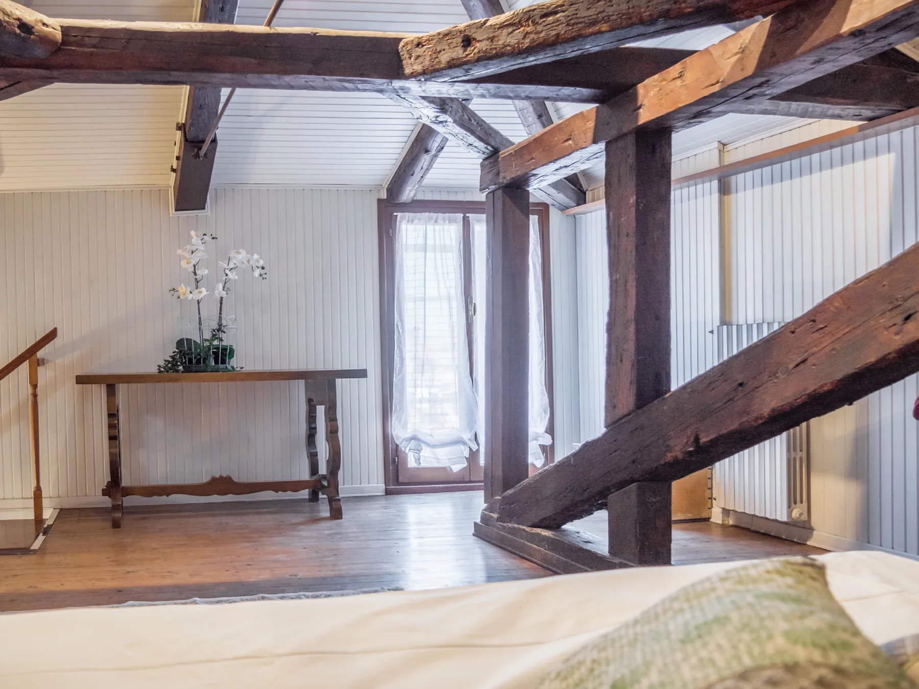 A bright attic room has exposed wooden beams and a bed in the foreground.