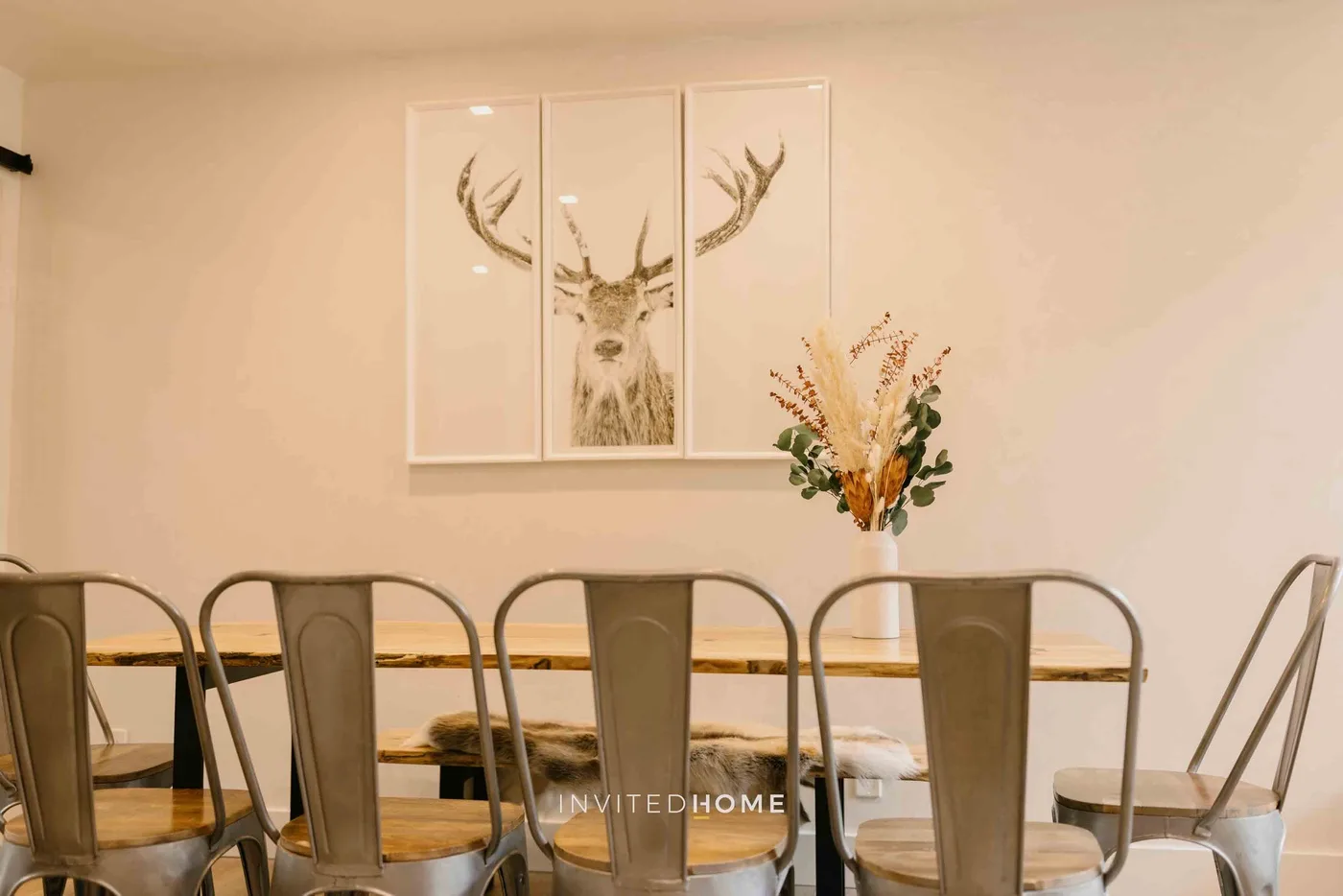 Un comedor con una mesa larga, asientos metálicos y arte enmarcado de un ciervo.