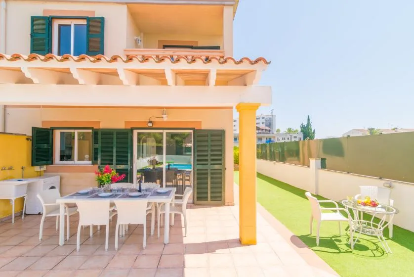 A sunny patio with outdoor dining tables beside a house and a strip of lawn.
