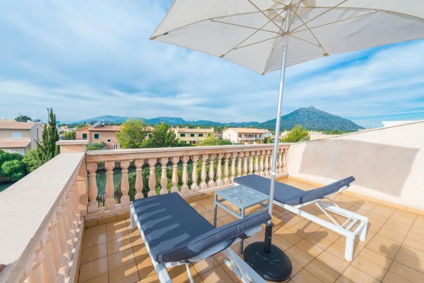 A sunny terrace has lounge seating beneath a large umbrella, with hills beyond.