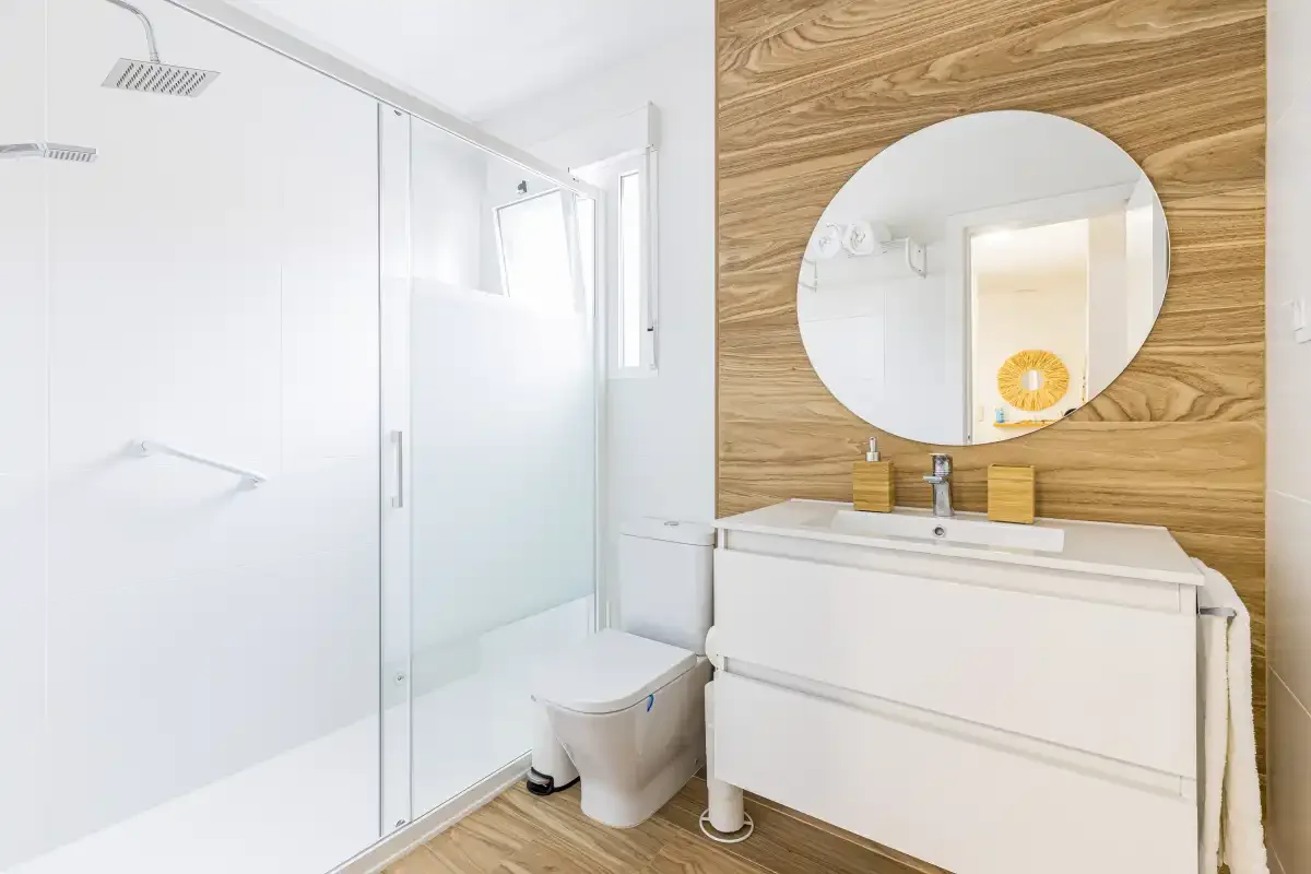 A bright bathroom with a glass shower and a white vanity beneath a round mirror.