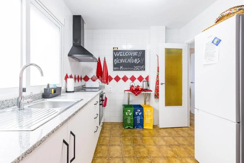 A bright tiled kitchen with a long countertop and colorful recycling bins.