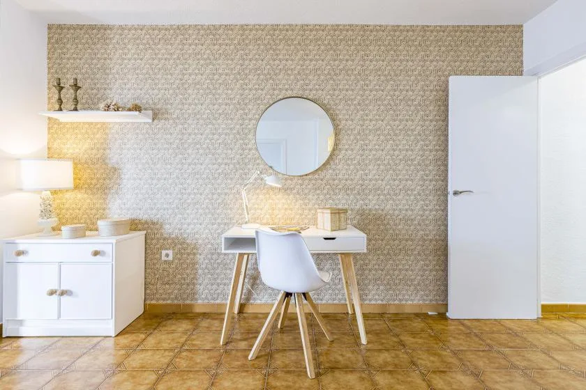 A bedroom vanity sits against a patterned wall, with a round mirror and chair.