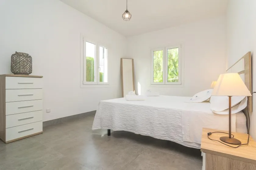 A bright bedroom with a bed, a dresser, and windows overlooking greenery.