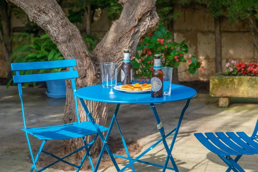 Una mesa azul de patio y asientos están bajo un árbol, junto a plantas con flores.