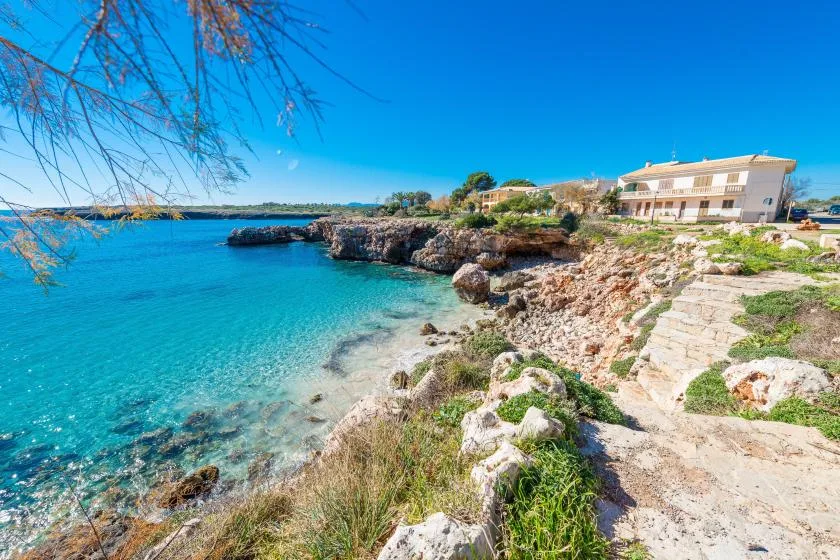 A rocky seaside landscape with clear blue water and a building along the shore.