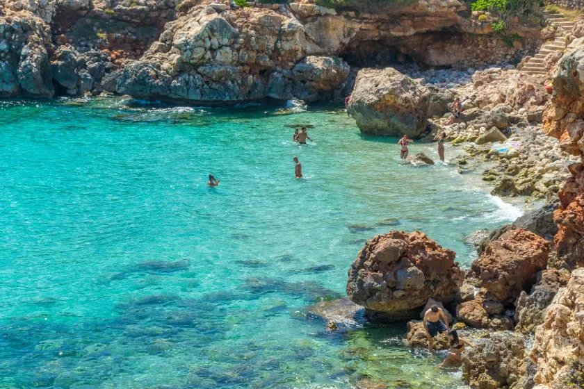 Clear turquoise water fills a rocky cove where people are swimming.