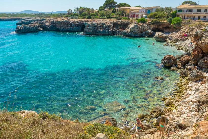 A rocky cove with clear turquoise water and buildings along the shore.