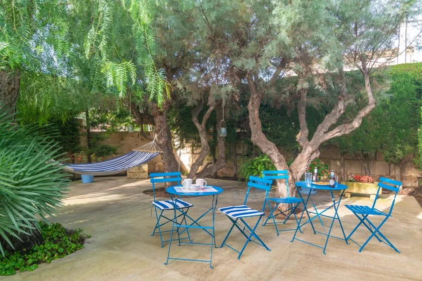 A shaded garden seating area has blue tables and chairs beneath leafy trees.