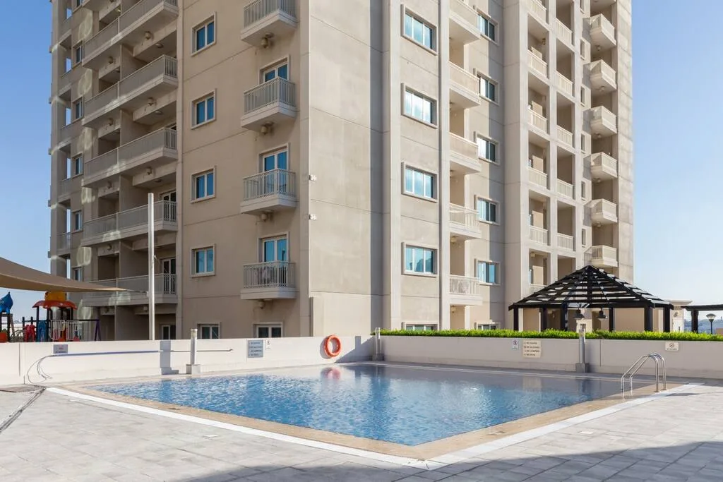 An outdoor swimming pool sits beside a high-rise residential building and shaded seating area.