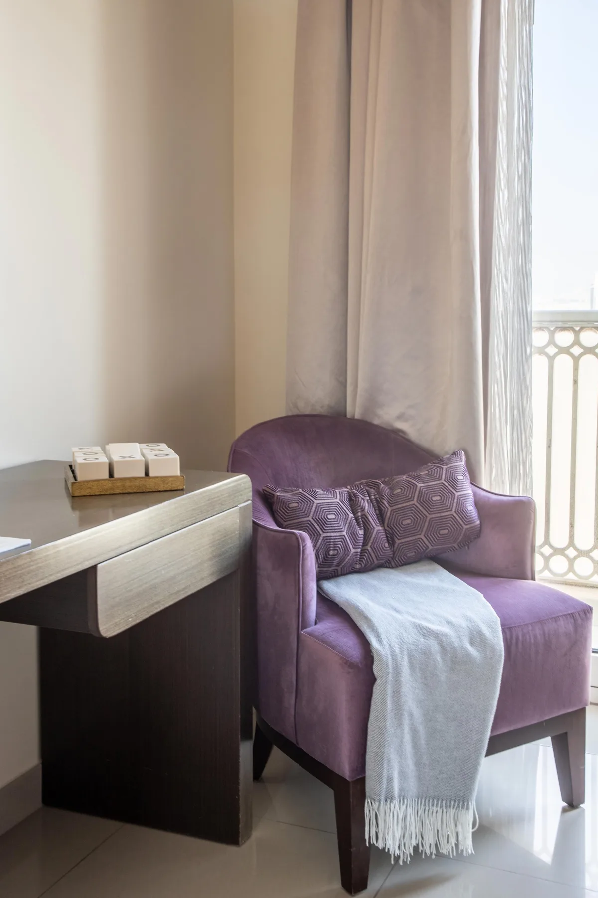 A purple armchair sits beside a desk near a curtained window.