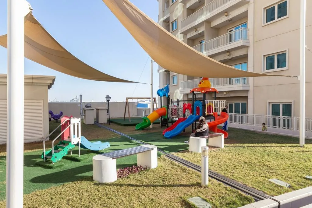 An outdoor playground with colorful slides sits beneath shade sails beside a building.