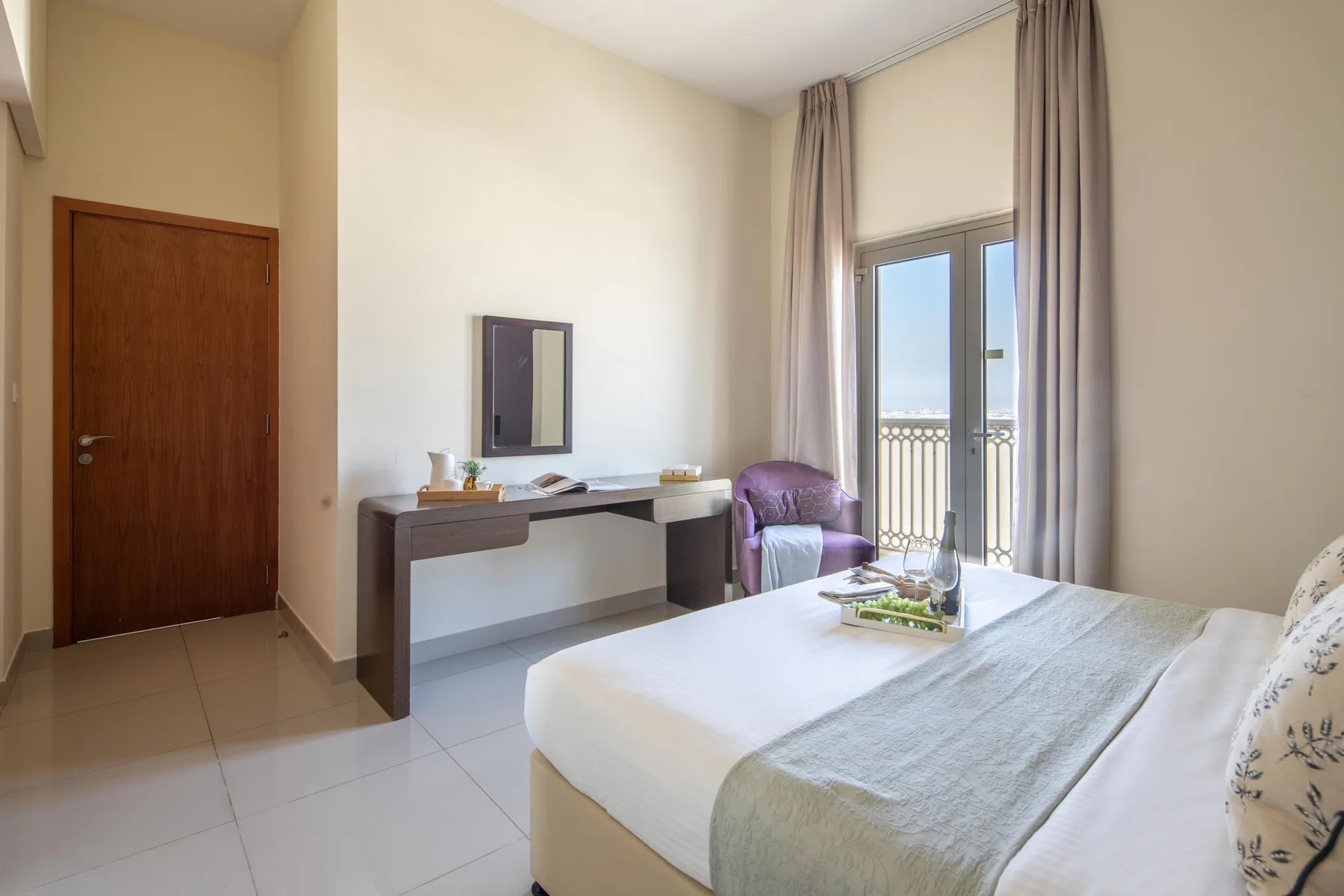 A bedroom with a neatly made bed, a desk, and glass doors opening onto a balcony.