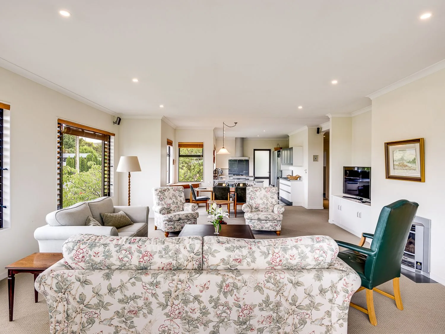 A living room with floral seating, a green armchair, a television, and an open kitchen area.