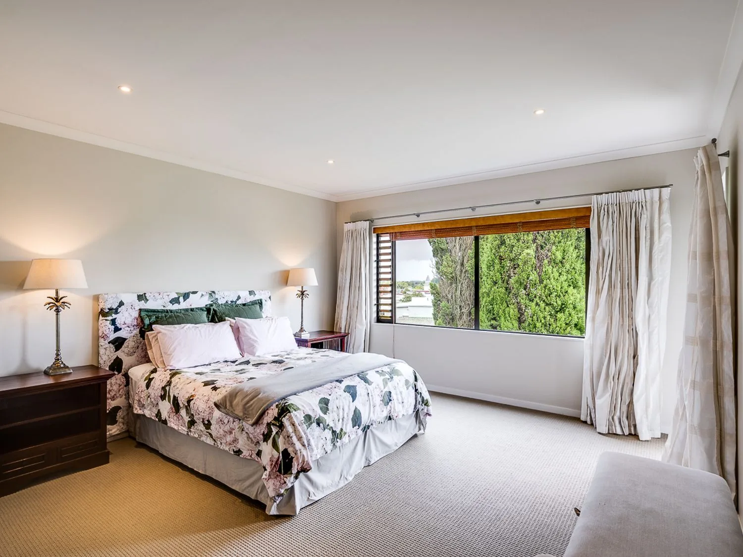 A bedroom with a bed with floral bedding two bedside tables with lamps a large window and curtains at Busby Hill Villa - Havelock North Holiday Home in Havelock North