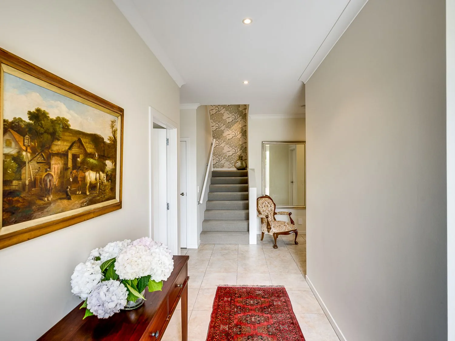 A hallway with a wooden table holding white flowers a painting on the left wall stairs straight ahead an upholstered chair and a mirror on the right at Busby Hill Villa - Havelock North Holiday Home in Havelock North