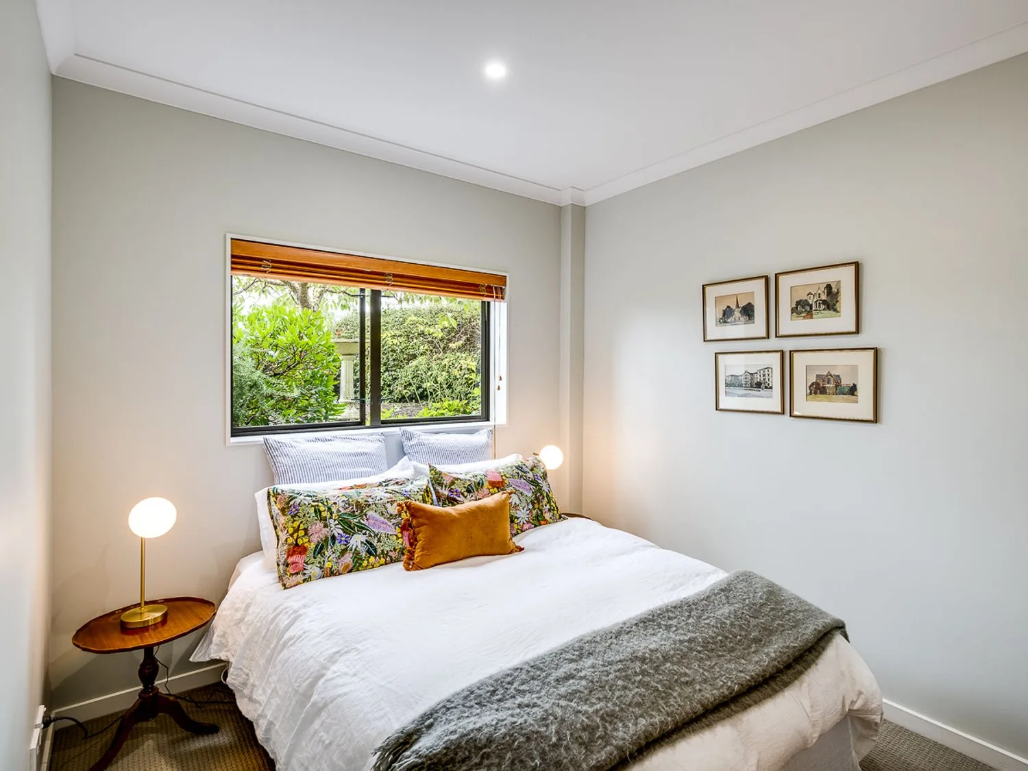 A bedroom with a bed with white bedding and floral pillows two bedside lamps a window and framed pictures on the wall at Busby Hill Villa in Havelock North