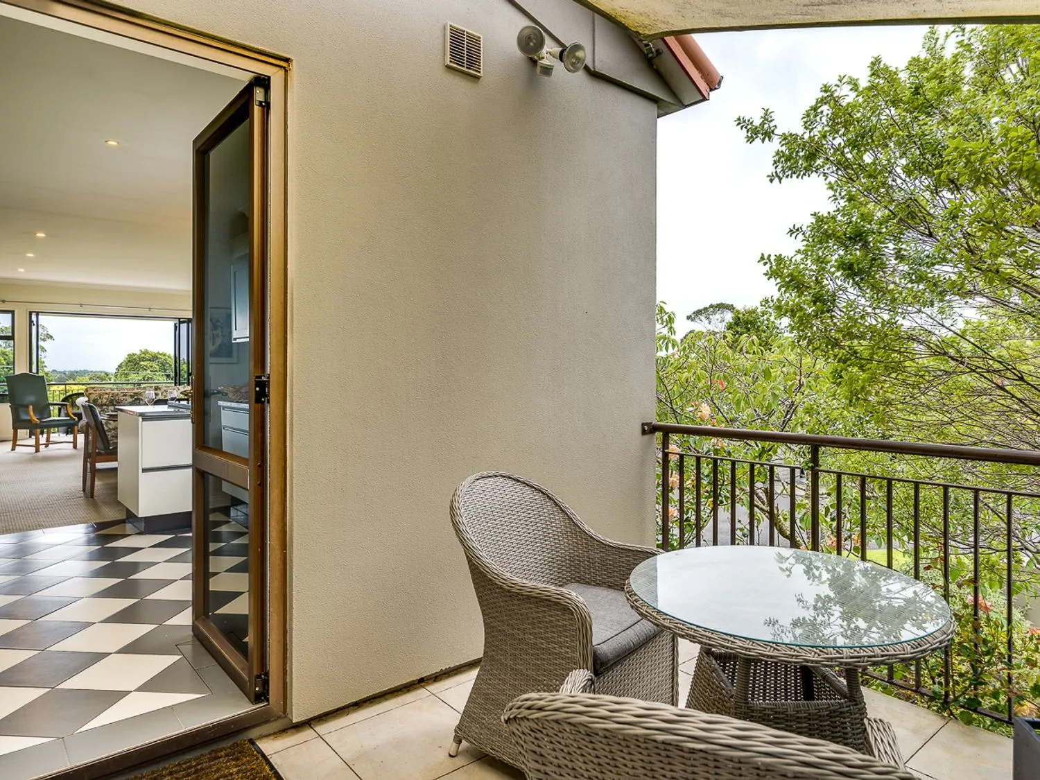A balcony with outdoor seating and a round glass-top table overlooks leafy trees.