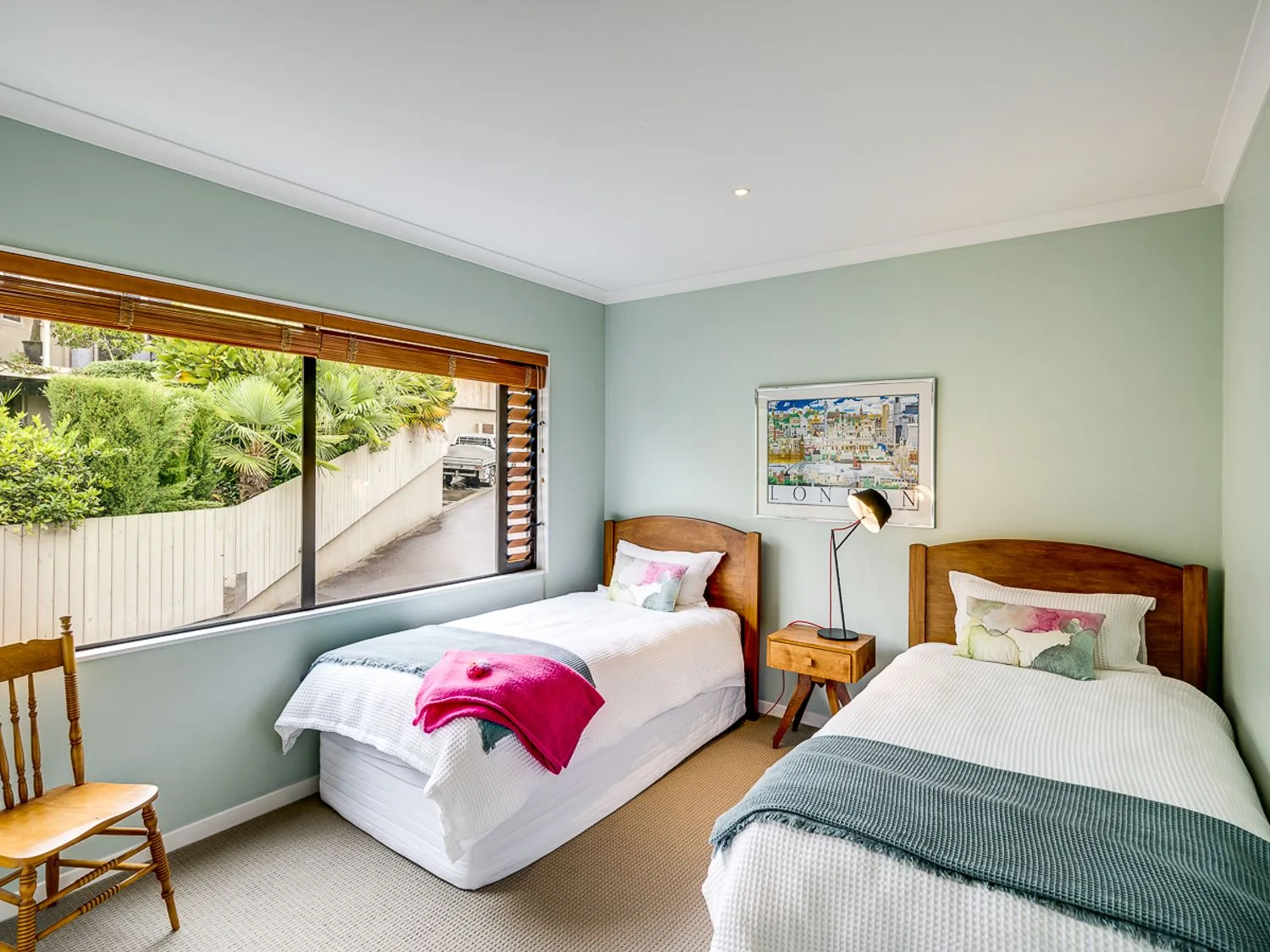 A bedroom with two single beds a wooden chair a bedside table with a lamp and a window showing outdoor greenery at Busby Hill Villa - Havelock North Holiday Home in Havelock North