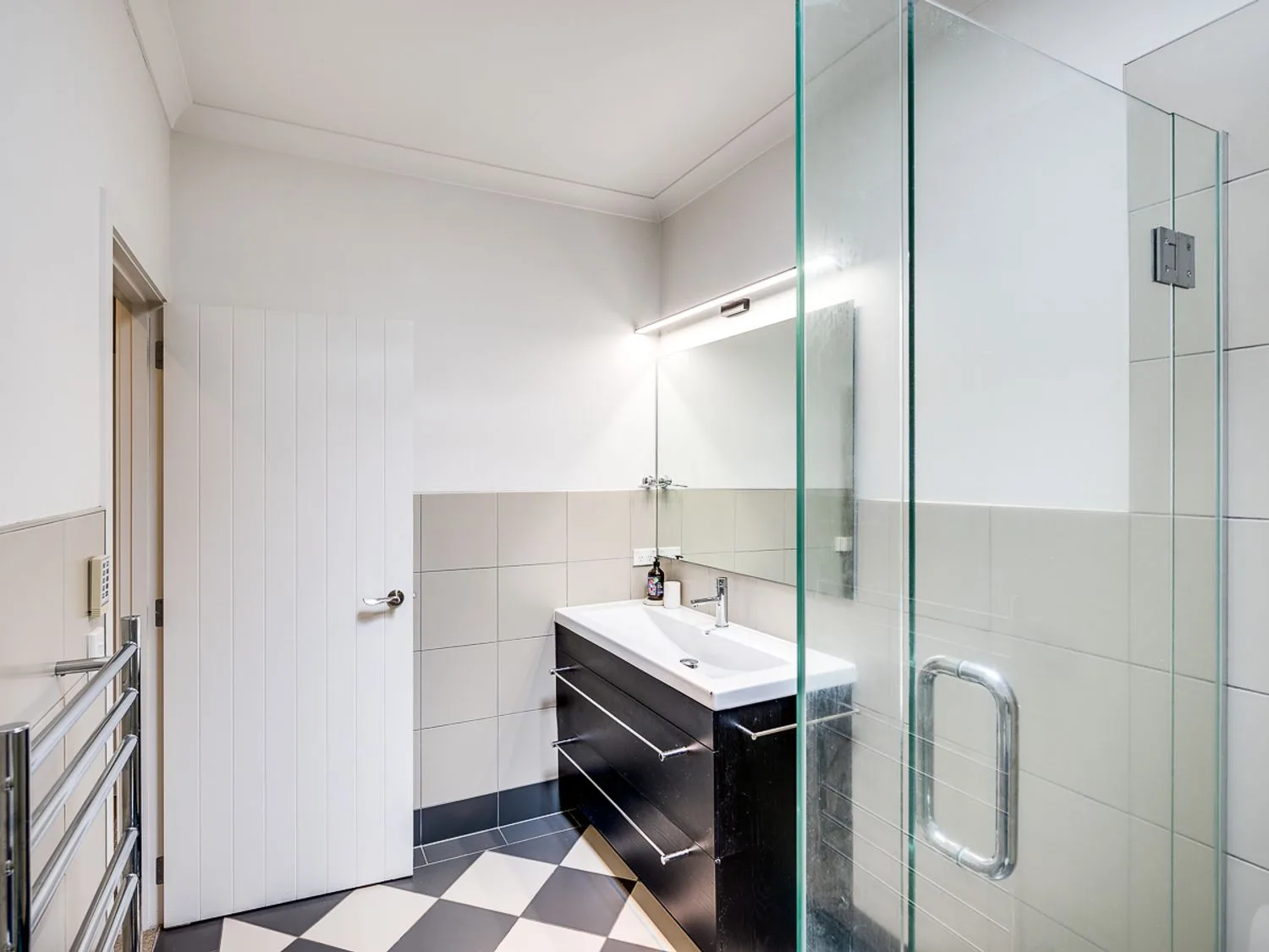A bathroom with a glass shower, a black vanity with a white sink, white tiled walls, and a metal towel rack at Busby Hill Villa in Havelock North