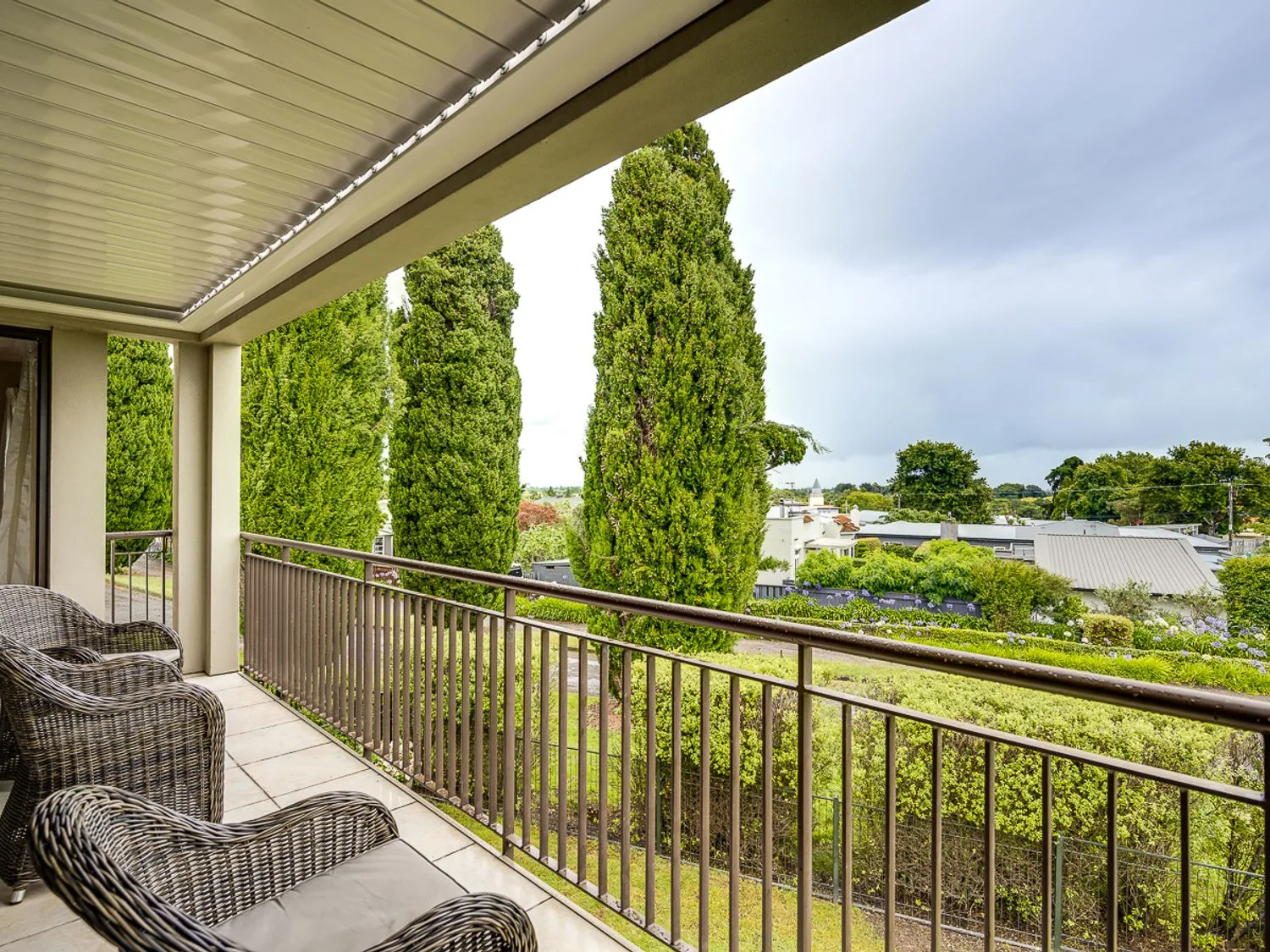 A balcony with wicker chairs and a railing overlooking trees and rooftops at Busby Hill Villa Havelock North