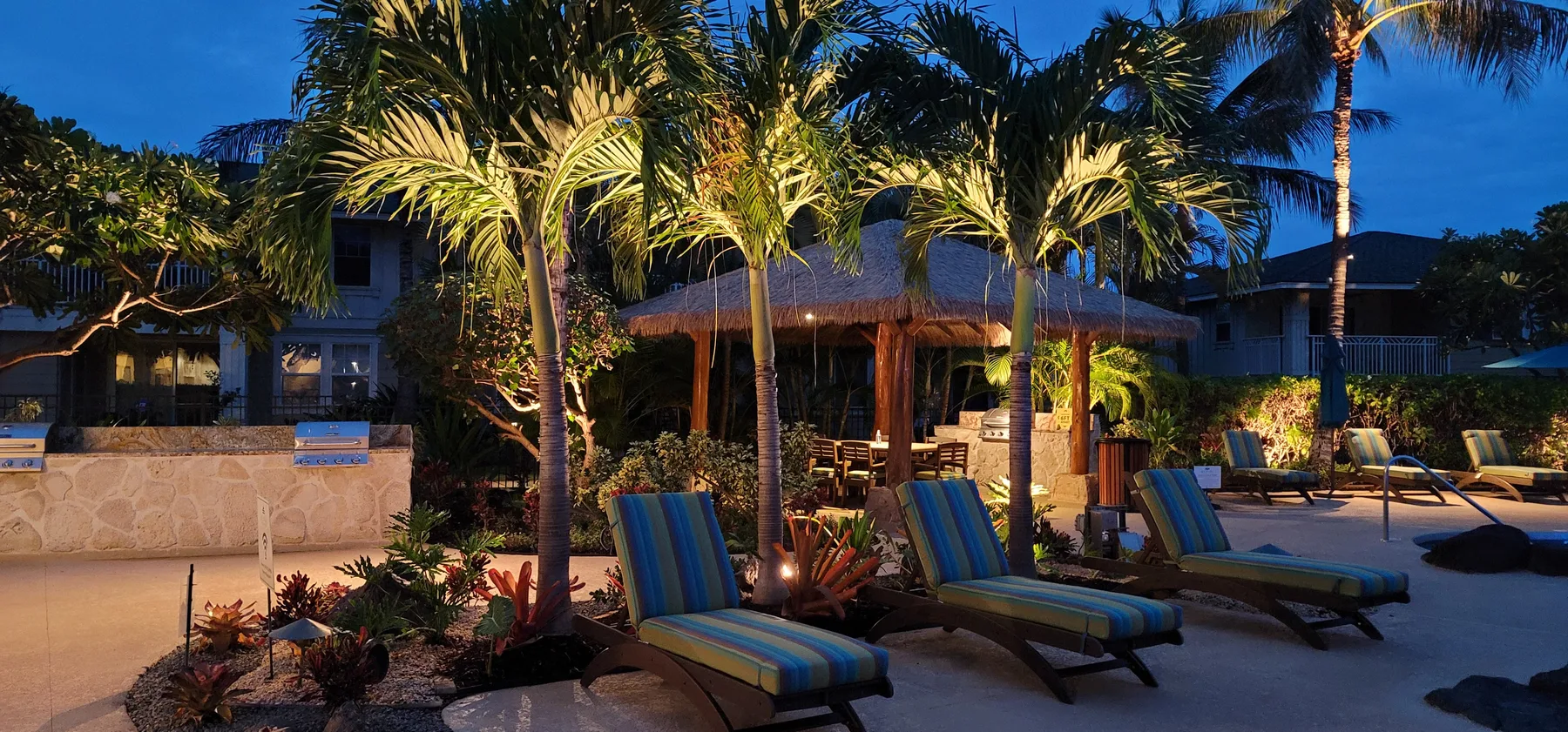 A poolside patio with striped lounge chairs beneath palm trees at dusk.