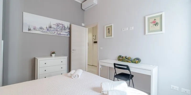 A bright bedroom with a bed, dresser, and desk against pale walls.